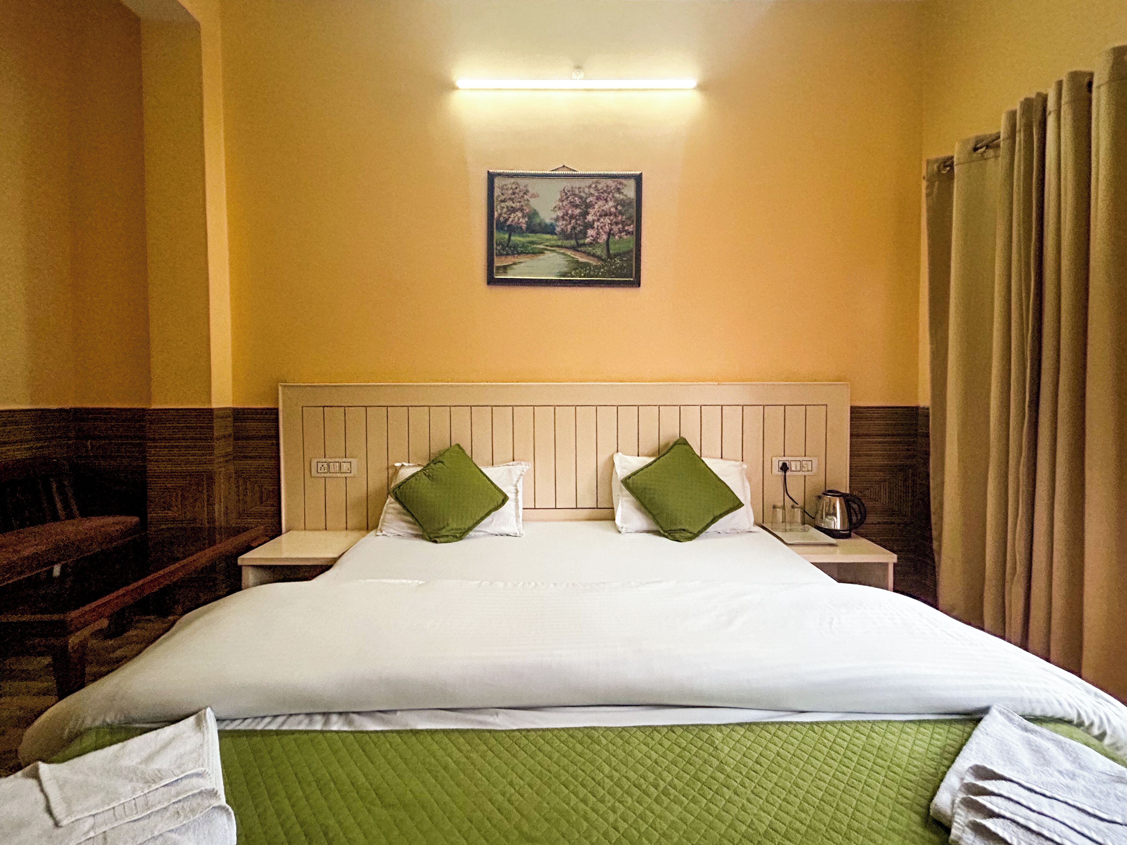 A hotel room with a bed, green pillows, a nightstand, a painting, and a window with curtains.