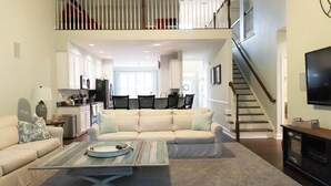 A spacious living room with a high ceiling, a staircase, and a kitchen in the background.