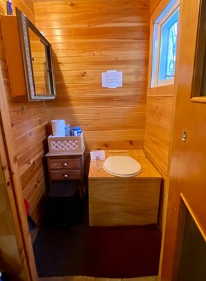 Ein kleines Badezimmer aus Holz mit Toilette, Spiegel, kleiner Ablage und Fenster.