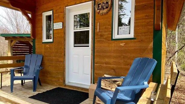 A wooden cabin with two blue Adirondack chairs on a wooden deck, a white door, and a sign that reads "ENZO".