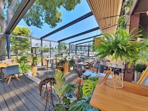 Un espacio exterior para comer con mesas y sillas, rodeado de plantas en macetas y vistas a un paisaje urbano.