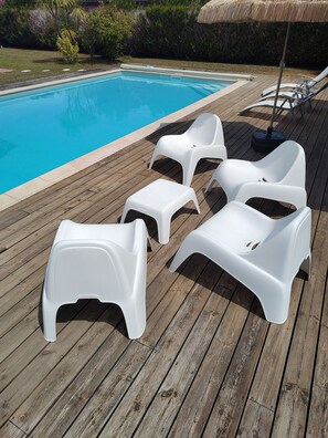 A wooden deck with a pool, white plastic lounge chairs, and a thatched umbrella.