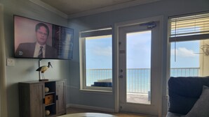 A living room with a glass coffee table, a television mounted on the wall, and a view of the ocean through a sliding glass door.