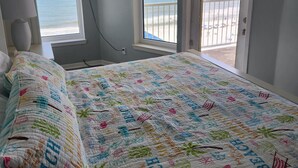 A bedroom with a bed, a ceiling fan, a TV mounted on the wall, and a view of the beach through two windows.
