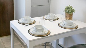 Ein weißer Esstisch mit vier Gedecken, jeweils bestehend aus Teller, Schüssel und Glas, auf gewebten Platzsets. Eine kleine Topfpflanze steht rechts auf dem Tisch.