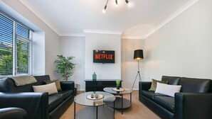 Ein modernes Wohnzimmer mit zwei schwarzen Ledersofas, einem Glastisch, einer schwarzen Stehlampe und einem an der Wand befestigten Fernseher, der "NETFLIX" zeigt.