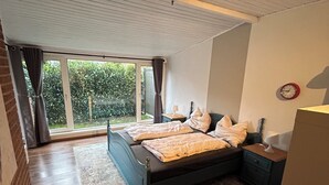 A bedroom with a large bed, a nightstand, a lamp, and a view of greenery through the window. - Schäferstübchen in Ekern near Bad Zwischenahn (Bad Zwischenahn)