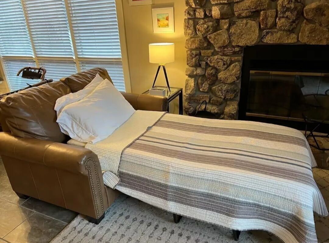 Una habitación acogedora con un sofá de cuero, una cama con manta a rayas, pared de piedra, chimenea y una lámpara.