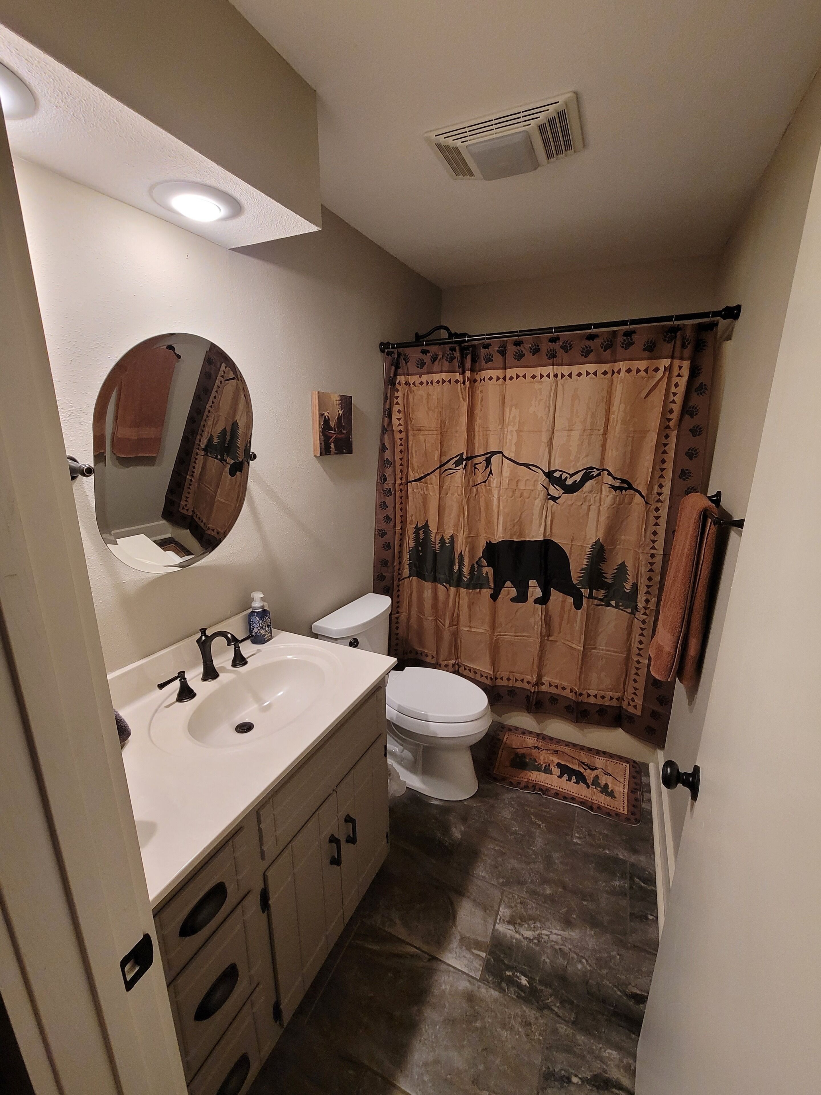 Un baño con lavamanos blanco, inodoro, espejo y cortina de ducha con diseño de bosque.