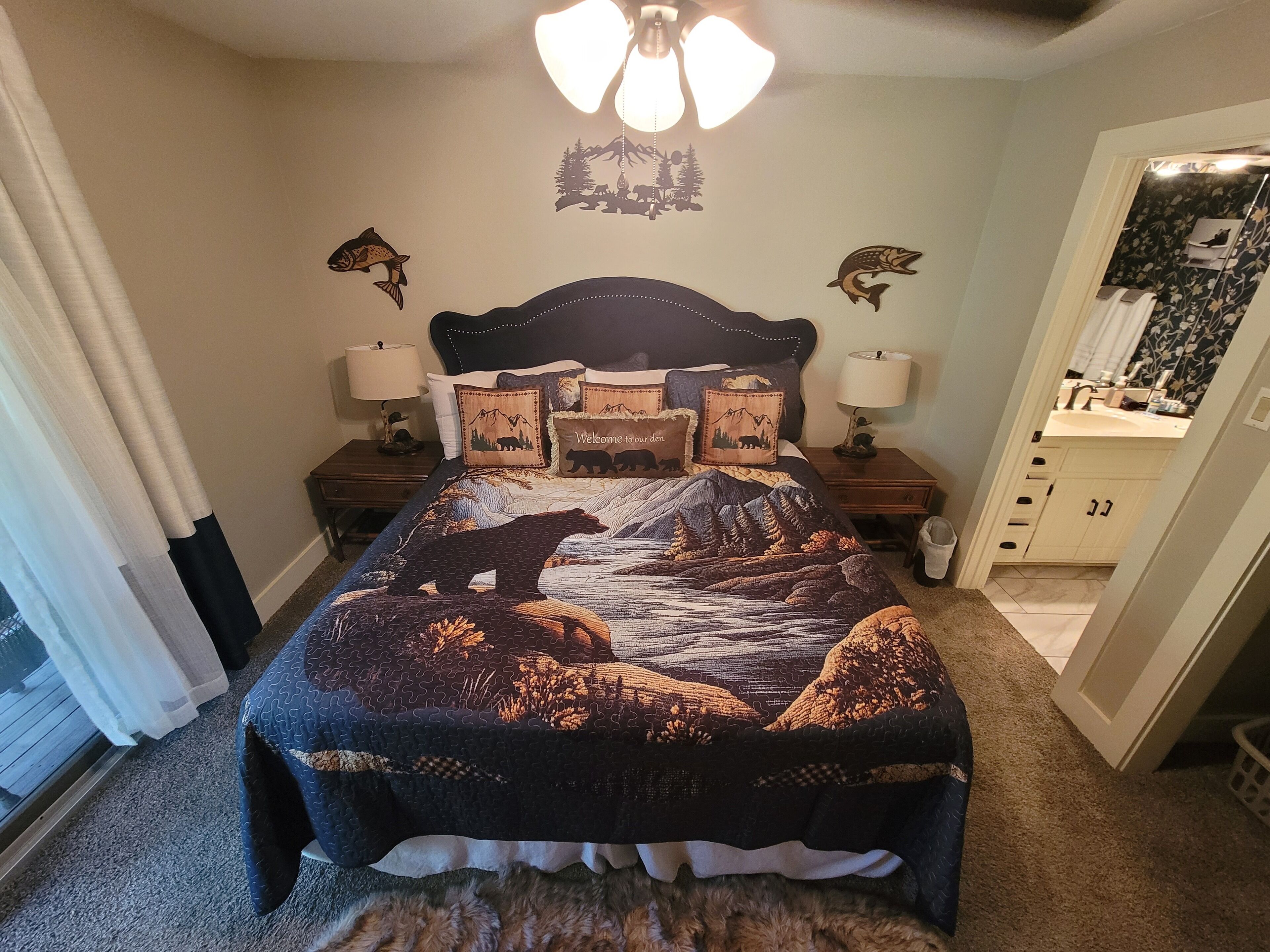 Un dormitorio con una cama que tiene un diseño de montaña y oso, dos mesitas de noche con lámparas, un tocador y un baño visible a través de una puerta abierta.