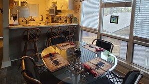 A dining area with a glass table and wooden chairs, a kitchen in the background, and a large window with blinds. - The Bear Den  (Mount Ida)