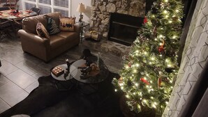 A cozy living room with a stone fireplace, a Christmas tree, a leather sofa, and a dining area with a table and chairs. - The Bear Den  (Mount Ida)