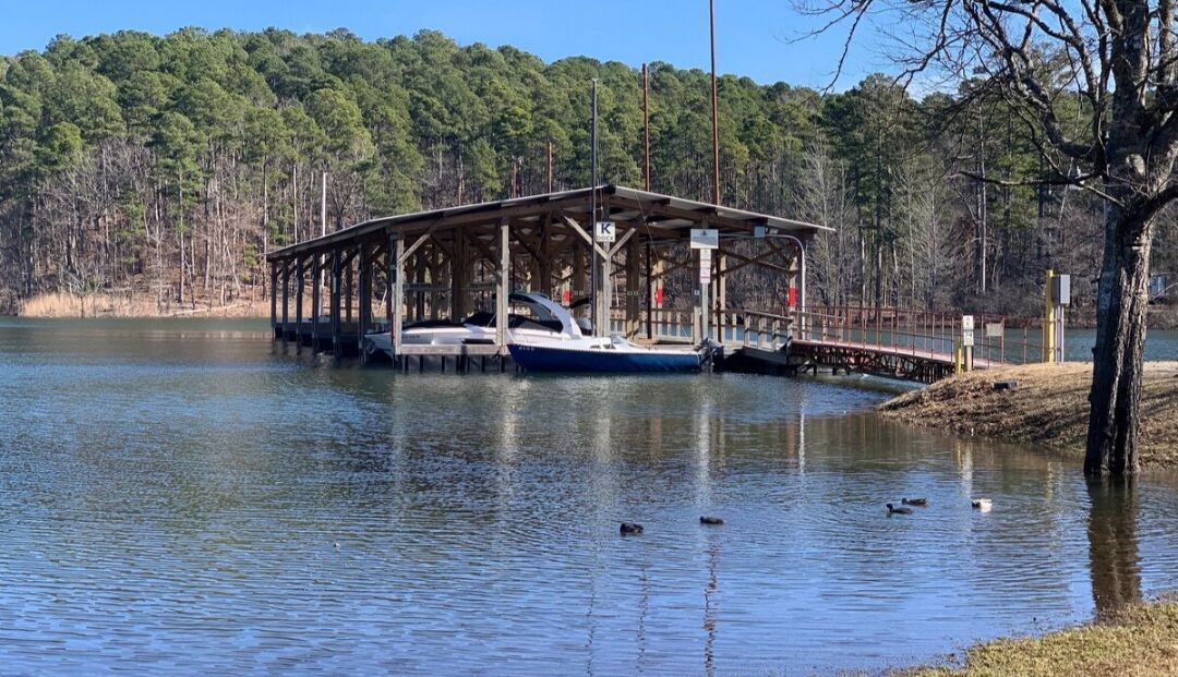 A docked boat at a pier with a covered shelter, surrounded by calm water and a forested area.