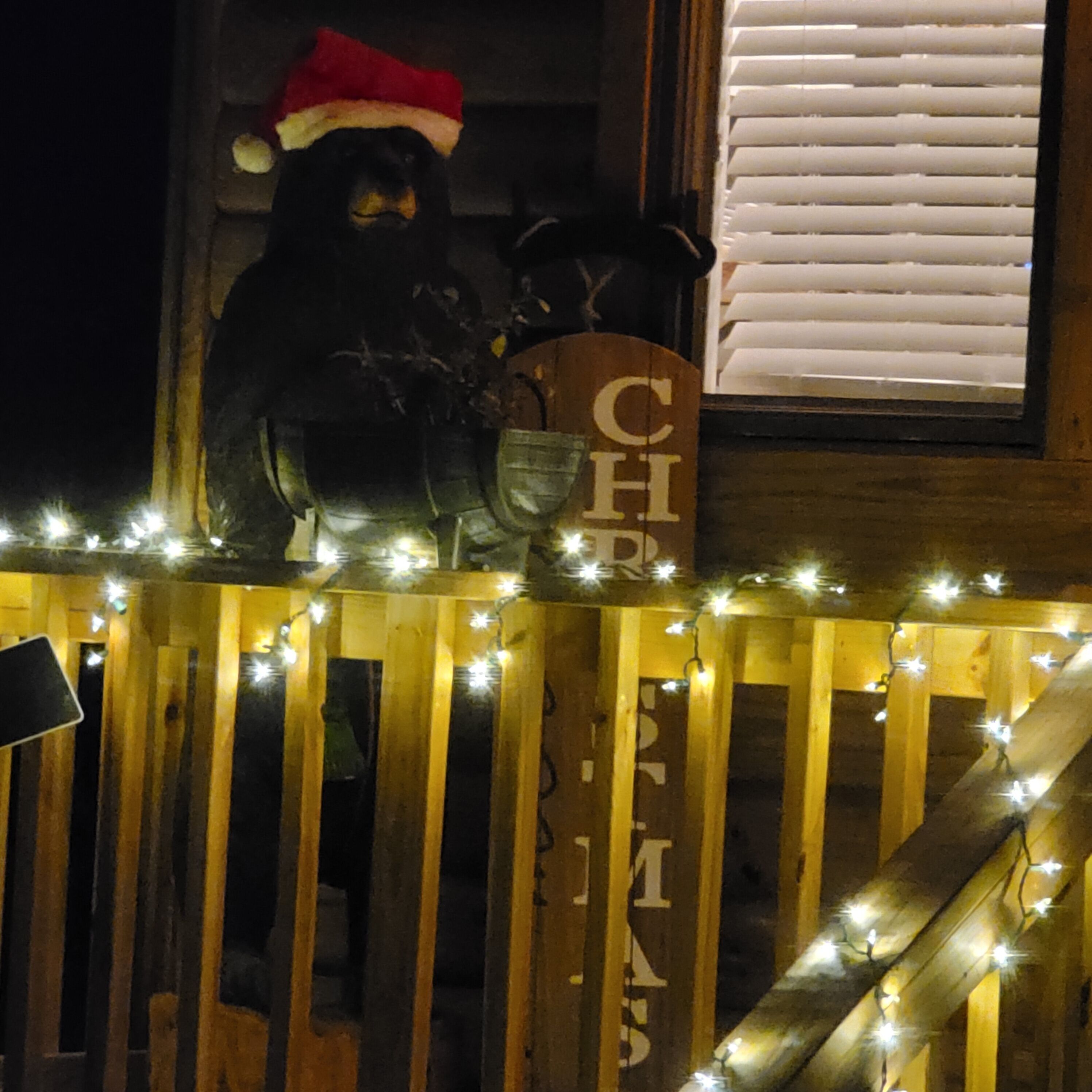 A wooden fence with a bear statue wearing a Santa hat, a sign with 'CHRISTMAS' written on it, and string lights.