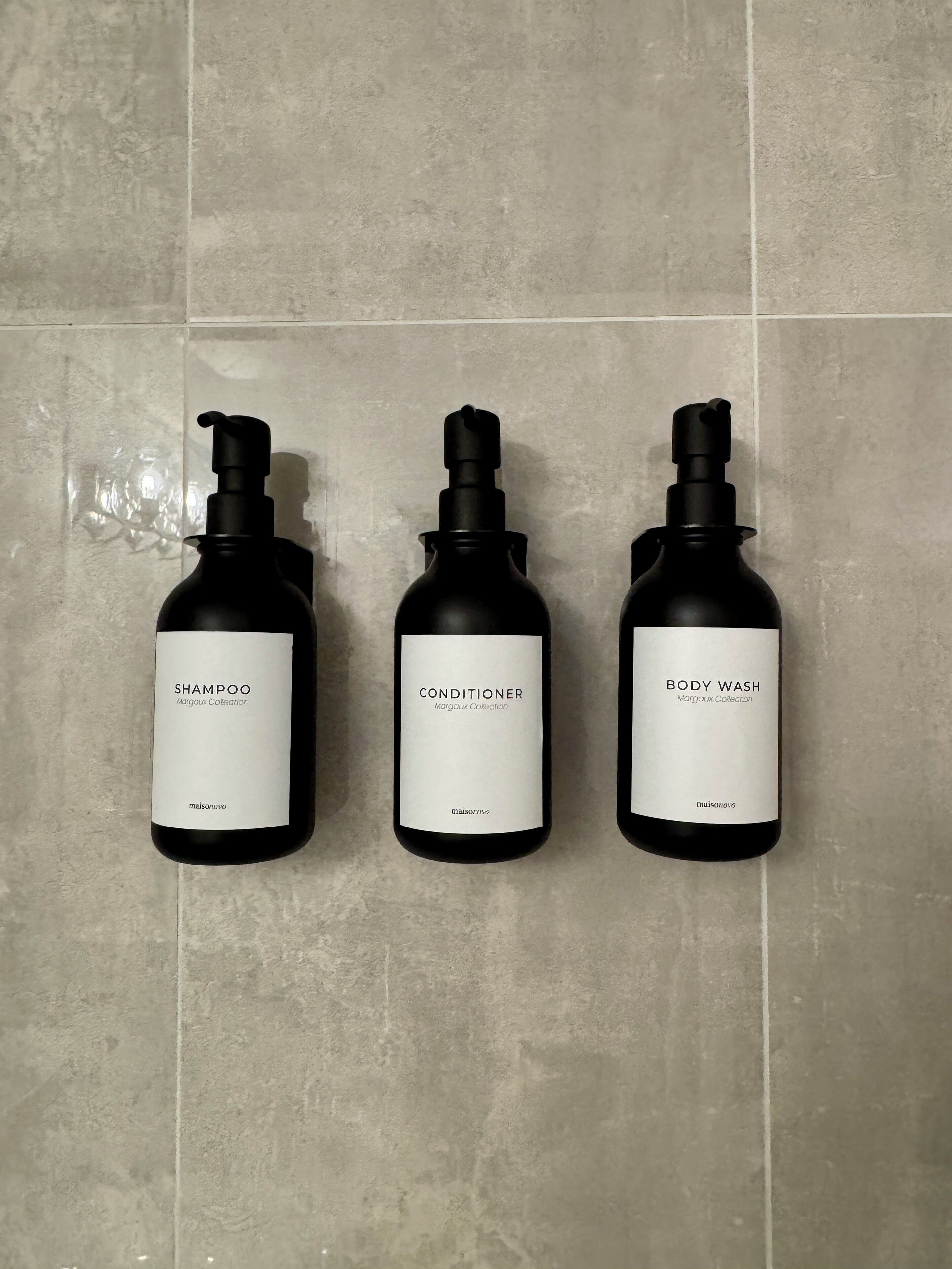 Three bottles of shampoo, conditioner, and body wash from the Margaux Collection, arranged on a tiled wall.