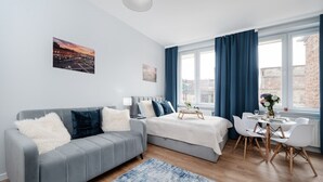 Ein modernes Schlafzimmer mit grauem Sofa, einem Bett mit Holzrahmen, einem Esstisch mit Stühlen und großen Fenstern mit blauen Vorhängen.
