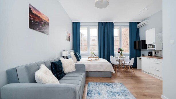 Ein modernes, gut beleuchtetes Zimmer mit einer grauen Couch, einem kleinen Esstisch und einer Küchenzeile.