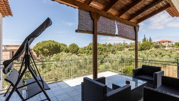 A covered outdoor patio with wicker furniture, a telescope, and a view of greenery and buildings in the background. - Lefkis apartments & studios (West Mani)