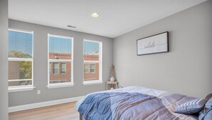 A bedroom with a large bed, three windows with blinds, a small bedside table, and a wall-mounted picture frame. - Spacious 3-bedroom apartment with AC, WiFi near Temple in marvelous Philadelphia (Philadelphia)