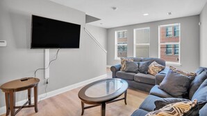A modern living room with a flat-screen TV mounted on the wall, a gray sofa with patterned cushions, a glass coffee table, and a wooden side table. - Spacious 3-bedroom apartment with AC, WiFi near Temple in marvelous Philadelphia (Philadelphia)