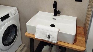 A bathroom with a white sink, a round mirror, a wooden shelf, and a washing machine. - Vacation home with panoramic sauna, free e-bike rental & space for 6 people! (Sankt Gertraud)