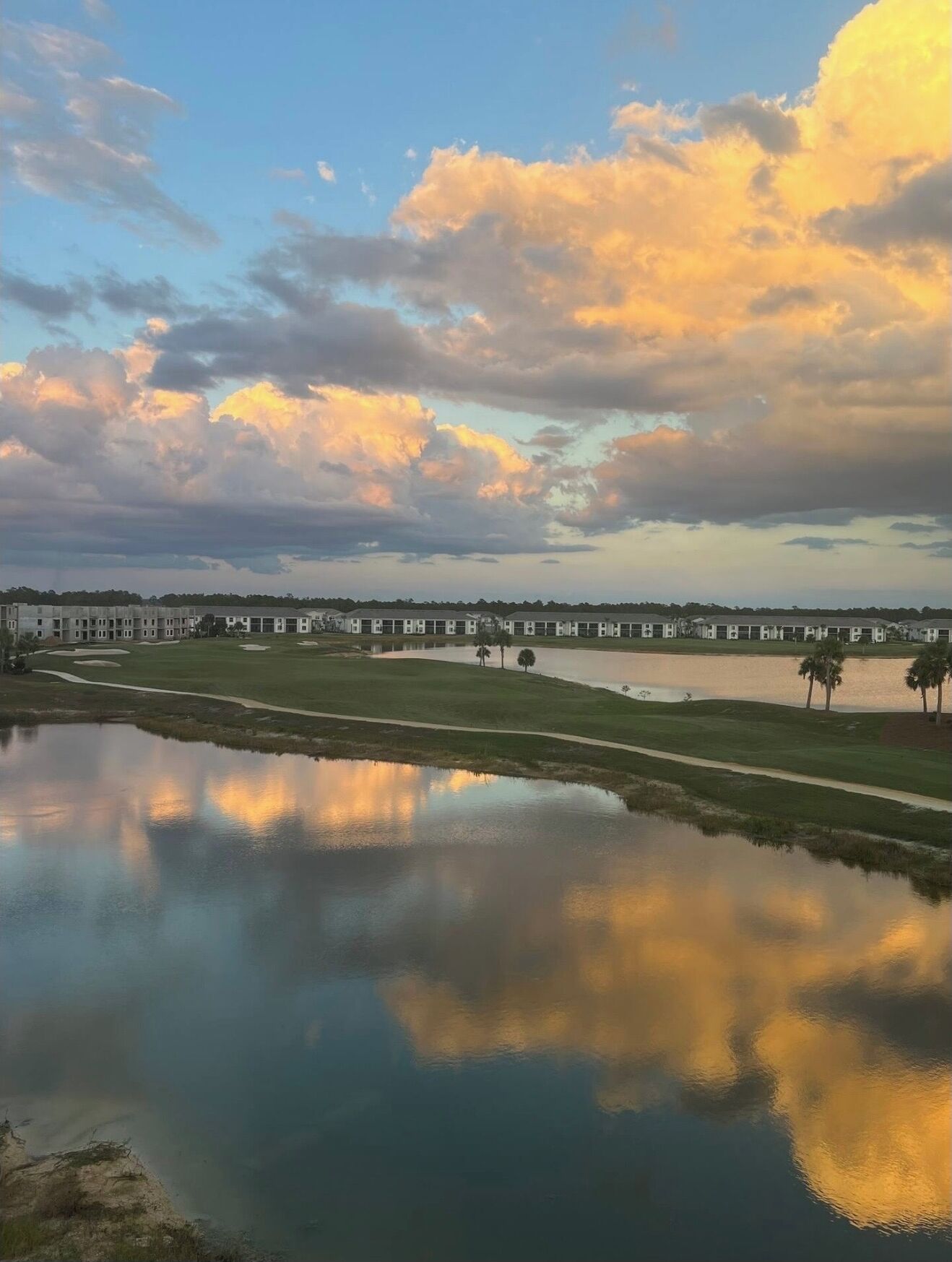 Un campo de golf con un cuerpo de agua que refleja el cielo, un puente y una fila de edificios al fondo.