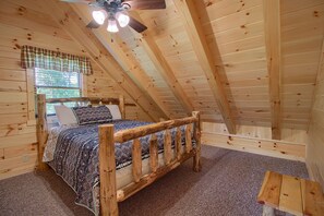 Interior de madeira com teto inclinado, uma cama com roupa de cama estampada e um pequeno banco de madeira.