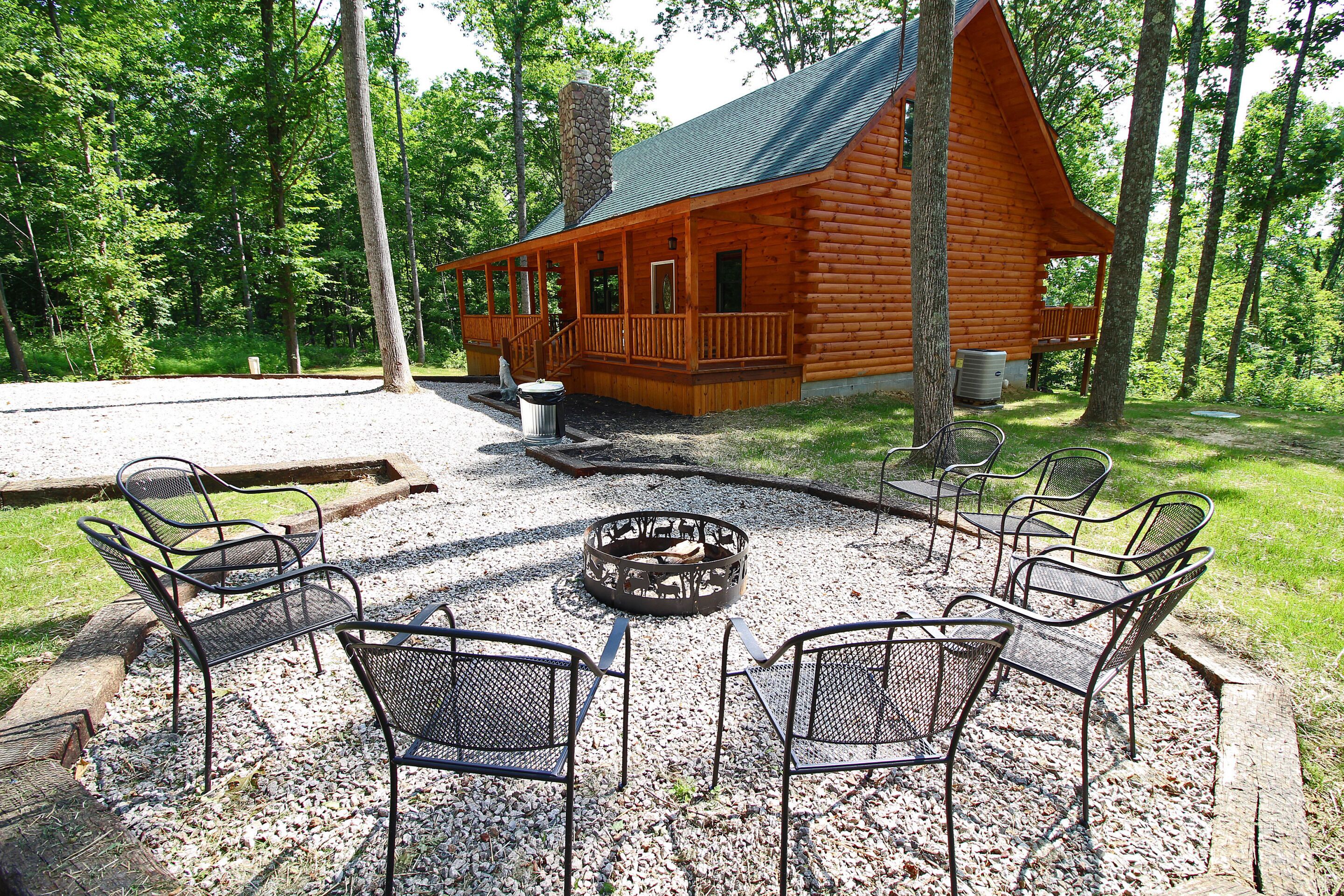 A log cabin with a porch, a fire pit, and metal chairs arranged around it.