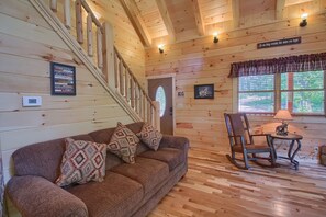 Interior de una cabaña con escalera de madera, sofá con cojines estampados, mecedora y una mesa con lámpara.