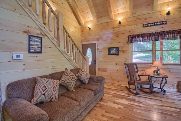 L’intérieur d’une cabane, avec un escalier en bois, un canapé agrémenté de coussins à motifs, un fauteuil à bascule et une table avec une lampe.