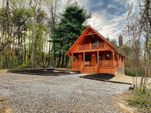 Une cabane en bois dotée d’un balcon, entourée d’une allée en gravier et d’une forêt.