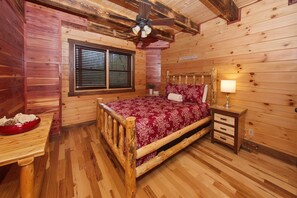 Une chambre dans une cabane en bois, comprenant un lit, une table de chevet, une lampe, une fenêtre avec des stores et un ventilateur de plafond.