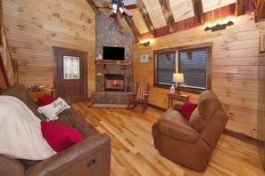 Een gezellig houten blokhutinterieur met een stenen openhaard, houten wanden en een comfortabele zithoek met leren banken en een schommelstoel.
