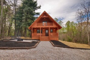 A wooden cabin with a balcony, surrounded by trees and a gravel driveway.