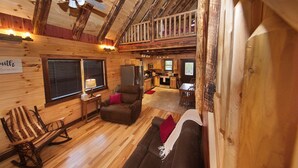 A rustic cabin interior with a wooden ceiling, a balcony, a kitchen area, and a cozy seating area with a rocking chair and sofa. - Kalli's Luxury Retreat- Unique Couple's Cabin (Logan)