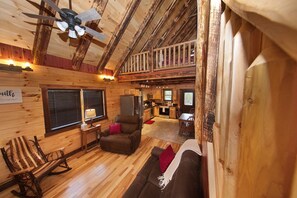 A rustic cabin interior with a wooden ceiling, a balcony, a kitchen area, and a cozy seating area with a rocking chair and sofa. - Kalli's Luxury Retreat- Unique Couple's Cabin (Logan)