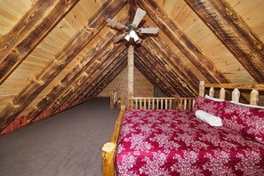 A wooden attic bedroom with a sloped ceiling, a wooden bed frame, a red floral bedspread, and a ceiling fan.