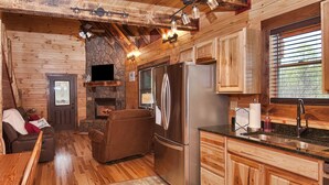 A rustic cabin interior with a stone fireplace, wooden walls, a large refrigerator, and a cozy seating area. - Kalli's Luxury Retreat- Unique Couple's Cabin (Logan)