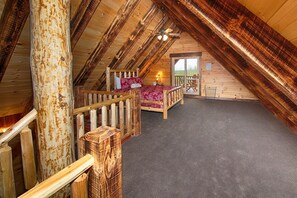 Een knus zolderkamer met een houten schuine dak, een bed met een rood geborduurde spreideken, en een klein balkon met een tafel en stoelen.