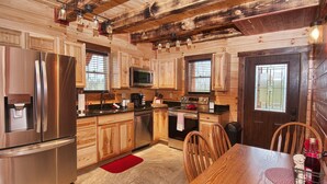 A modern kitchen with wooden cabinetry, stainless steel appliances, and a dining area with a wooden table and chairs. - Kalli's Luxury Retreat- Unique Couple's Cabin (Logan)