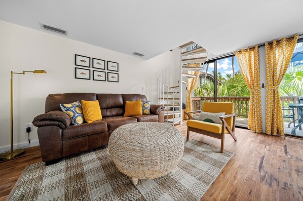 Ein Wohnzimmer mit einem braunen Ledersofa, gelben Kissen, einem runden gewebten Hocker, einer Stehlampe und Blick auf einen Balkon mit gelben Vorhängen.