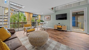 Un espacio habitable amplio con piso de madera, un sofá marrón, un otomán tejido y un comedor. Un televisor está montado en la pared, y una puerta corrediza da a un área exterior con vista a palmeras.