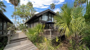 Una terraza de madera que conduce a una casa con balcón, rodeada de vegetación tropical.