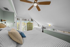 Ein Schlafzimmer mit schrägem Dach, Deckenventilator, Bett mit weißer Bettwäsche und Blick ins Freie durch ein Fenster mit weißen Jalousien.
