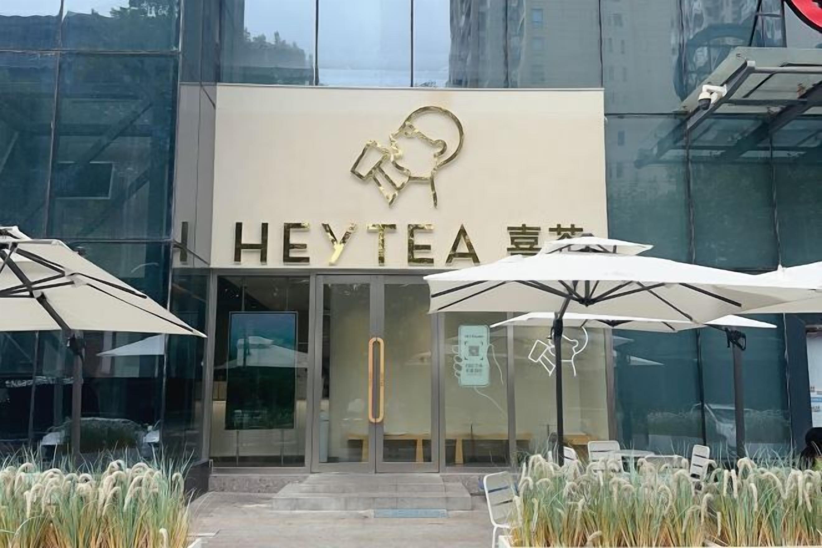 Un bâtiment moderne avec une façade en verre et une enseigne indiquant « HEYTEA », sur laquelle figure un logo. Devant l’entrée, on trouve des parasols et des espaces aménagés pour s’asseoir.