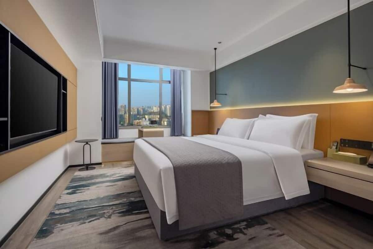 Quarto de hotel moderno com uma cama grande, uma televisão, vista para a cidade e uma mesa pequena.