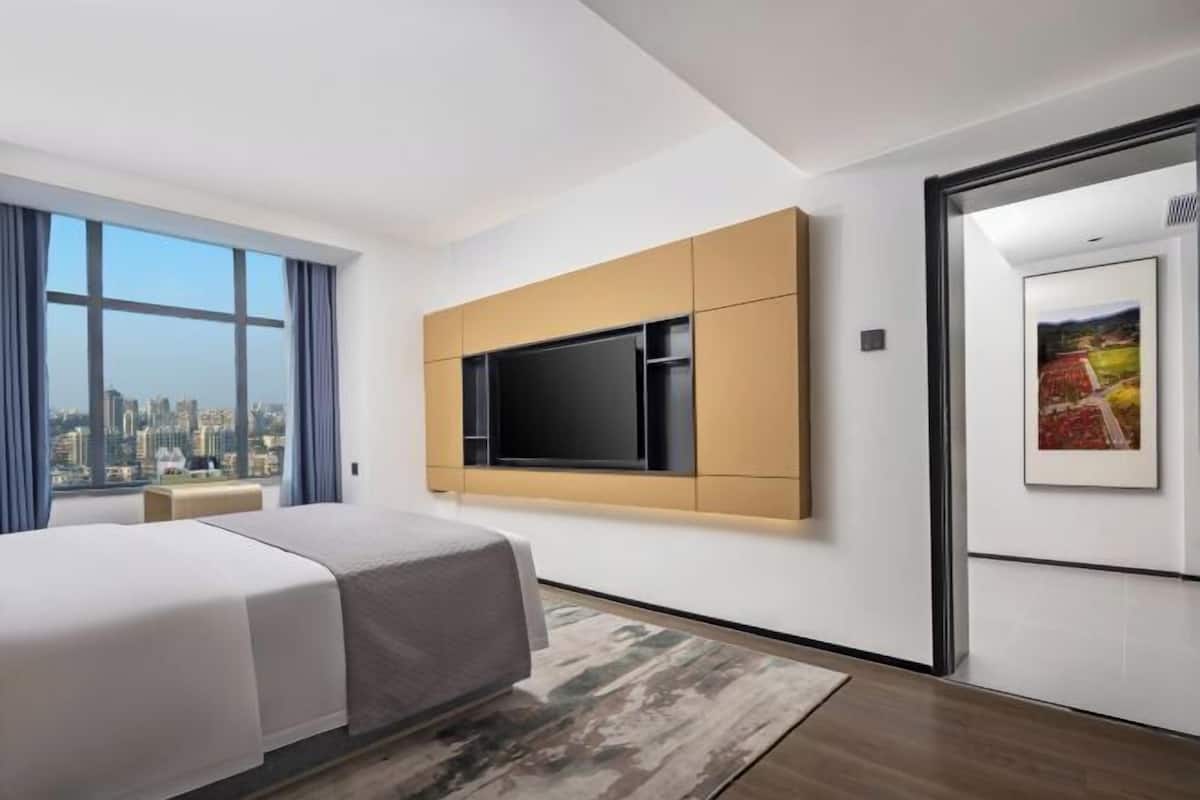 Quarto de hotel moderno com cama, uma grande janela com vista para a cidade, televisão montada e um quadro na parede.