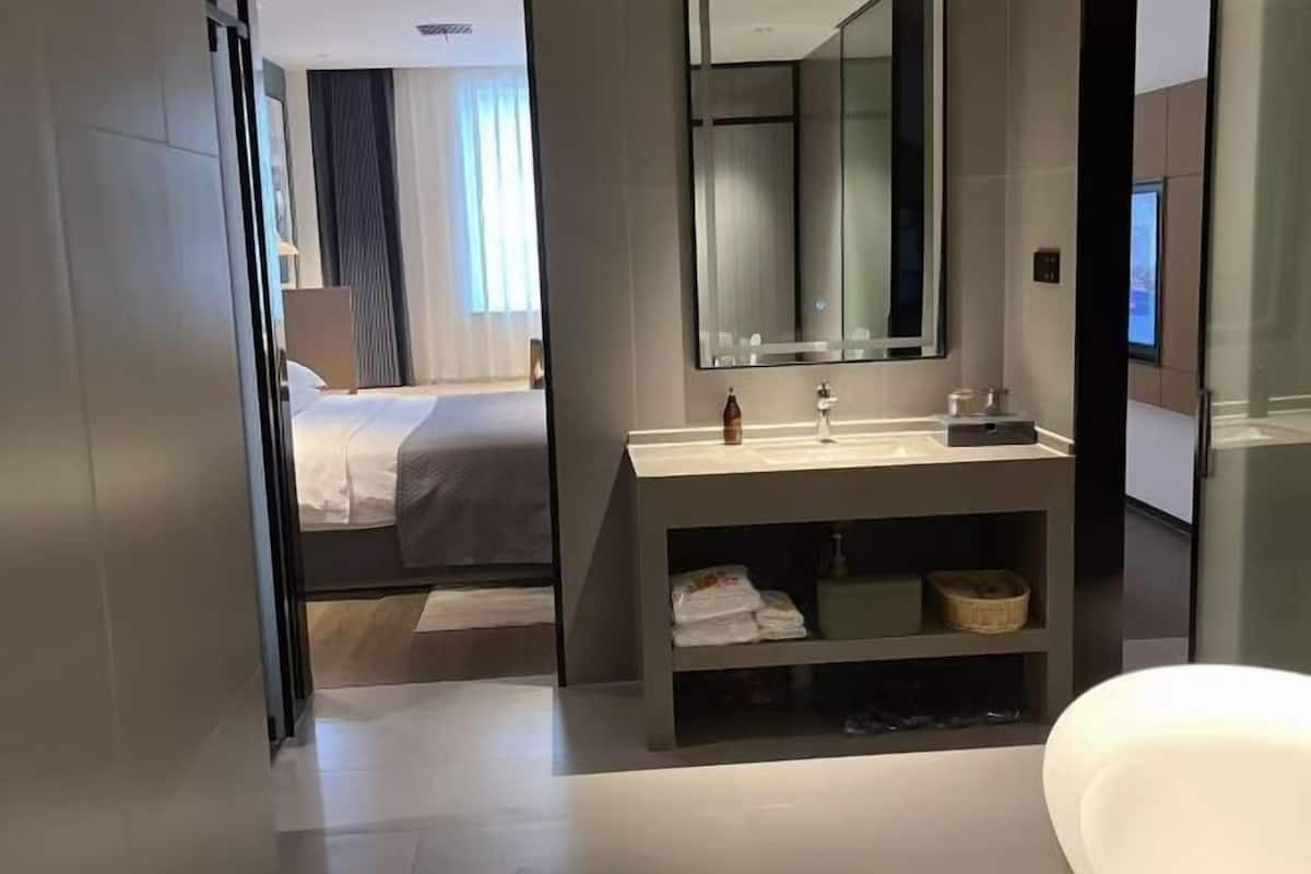 Quarto de hotel moderno com uma cama grande, banheiro com toucador e espelho, e vista da cidade pela janela.