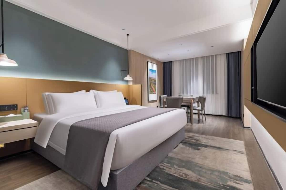 Quarto de hotel com cama, mesa de cabeceira, luminária, escrivaninha, cadeira e uma grande janela com cortinas.