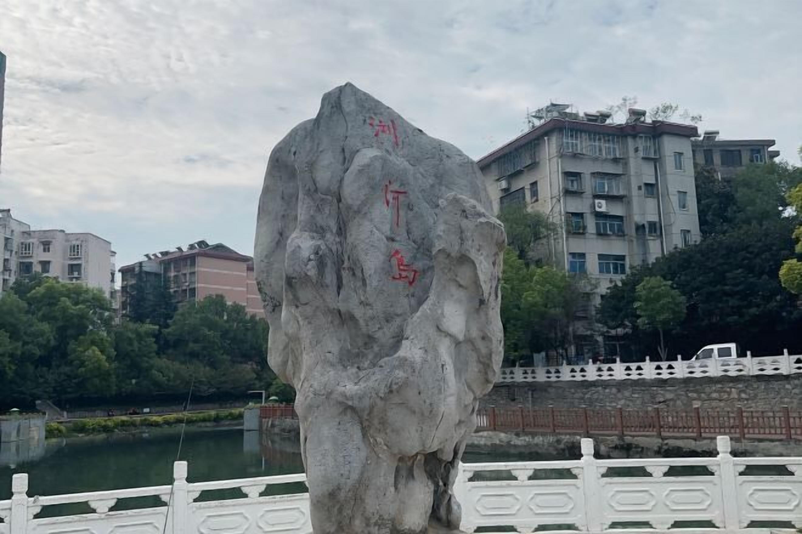 A large rock with inscriptions, surrounded by a white fence and buildings in the background.